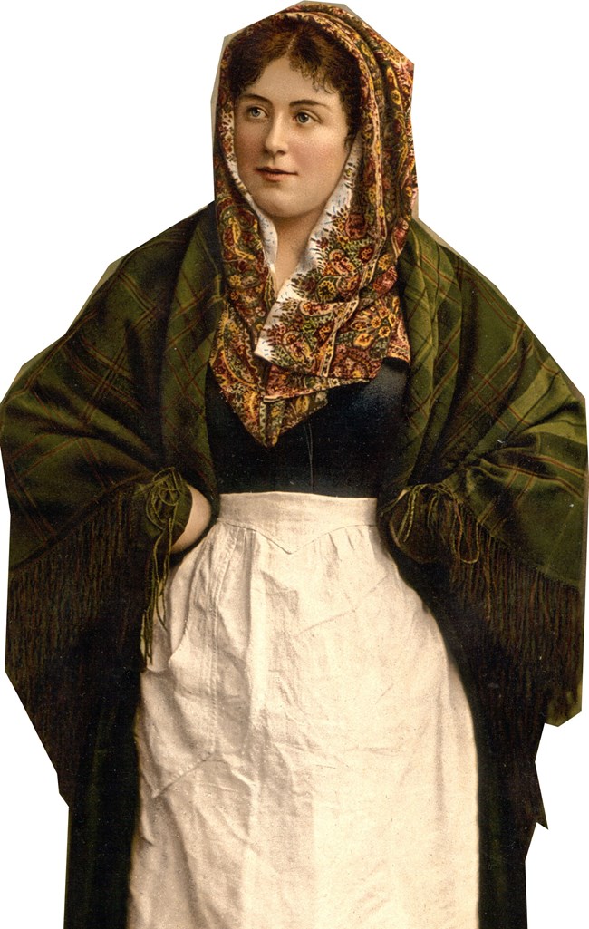 A woman with a head scarf and a brown/green shawl that covers her black blouse and white dress