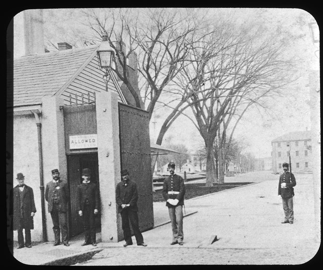 Photograph of men in uniforms and suits by a entrance to a roadway. A gas lamp is above a post for the gate.