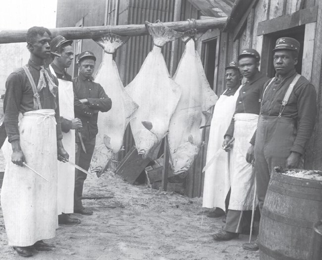 six black men wearing military uniforms and aprons stand alongside three enormous fish strung on a pole.
