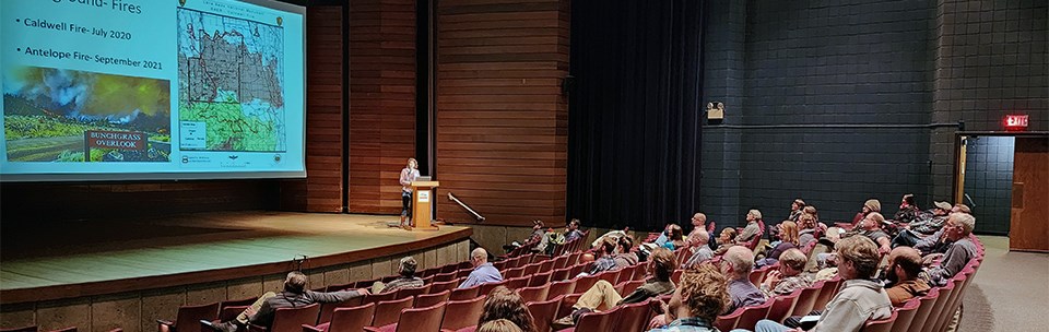 A scattered group sits in an auditorium looking at a speaker and screen on the stage. Screen has map and information about wildfires at Lava Beds National Monument.