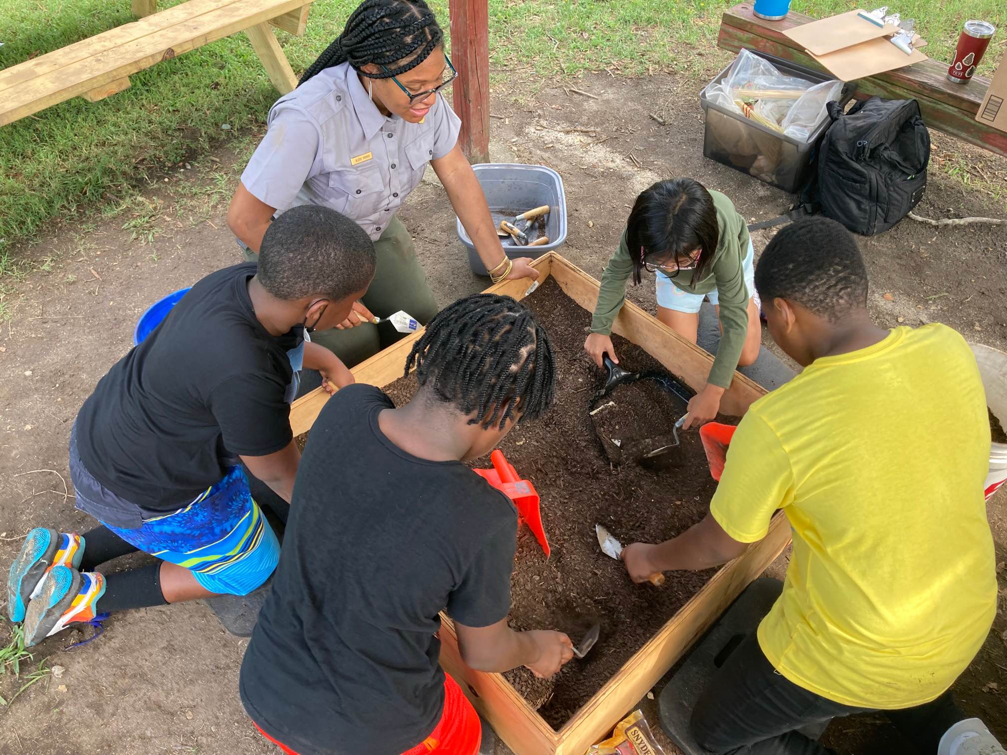 A uniformed NPS ranger and four kids excavate with trowels and scoopers in a dig box