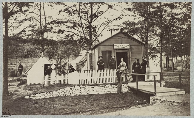 Black and white photograph of wood building with U.S. Sanitary Commission on the front. There are white camping tents next to the building, and, spread out, men wearing a dark soldiers uniform. There are two women in dresses standing in front of the build