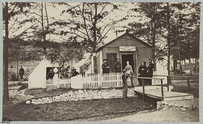 convalescentcamp Black and white photograph of wood building with U.S. Sanitary Commission on the front. There are white camping tents next to the building, and, spread out, men wearing a dark soldiers uniform. There are two women in dresses standing in front of the build