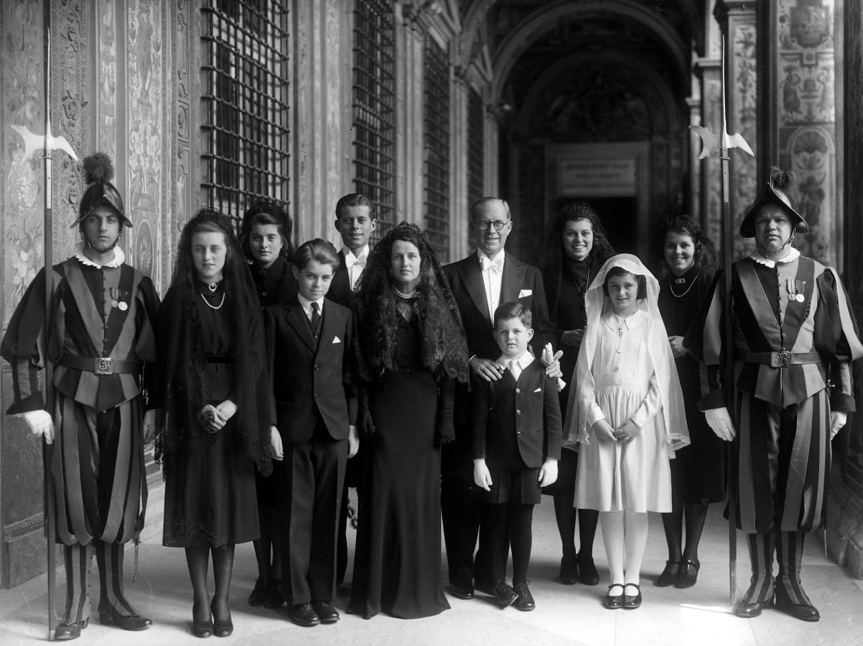 A black and white photo of a family of ten standing in an arched walkway, between two Swiss Guards in striped uniforms with halberds. The men and boys wear black suits, while the women and girls wear black dresses and veils, except the youngest, in white.