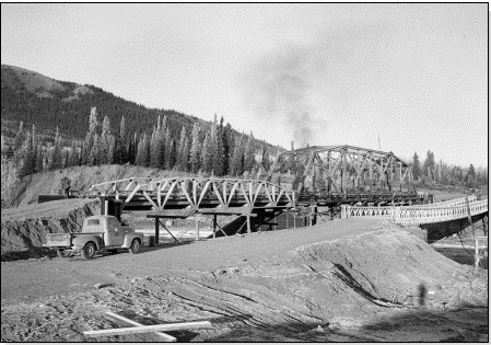 A black and white photo of a bridge under construction. Scaffolding supports the ends of the bridge and dirt roads ramp up to where the bridge ends.