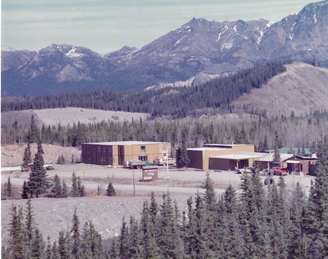 A few buildings sit on the edge of clearing in the forest. A few trucks and vans are parked on the edge of an empty parking lot. A brown sign reads "Mt McKinley Village". Rugged, rocky mountains rise up in the distance.