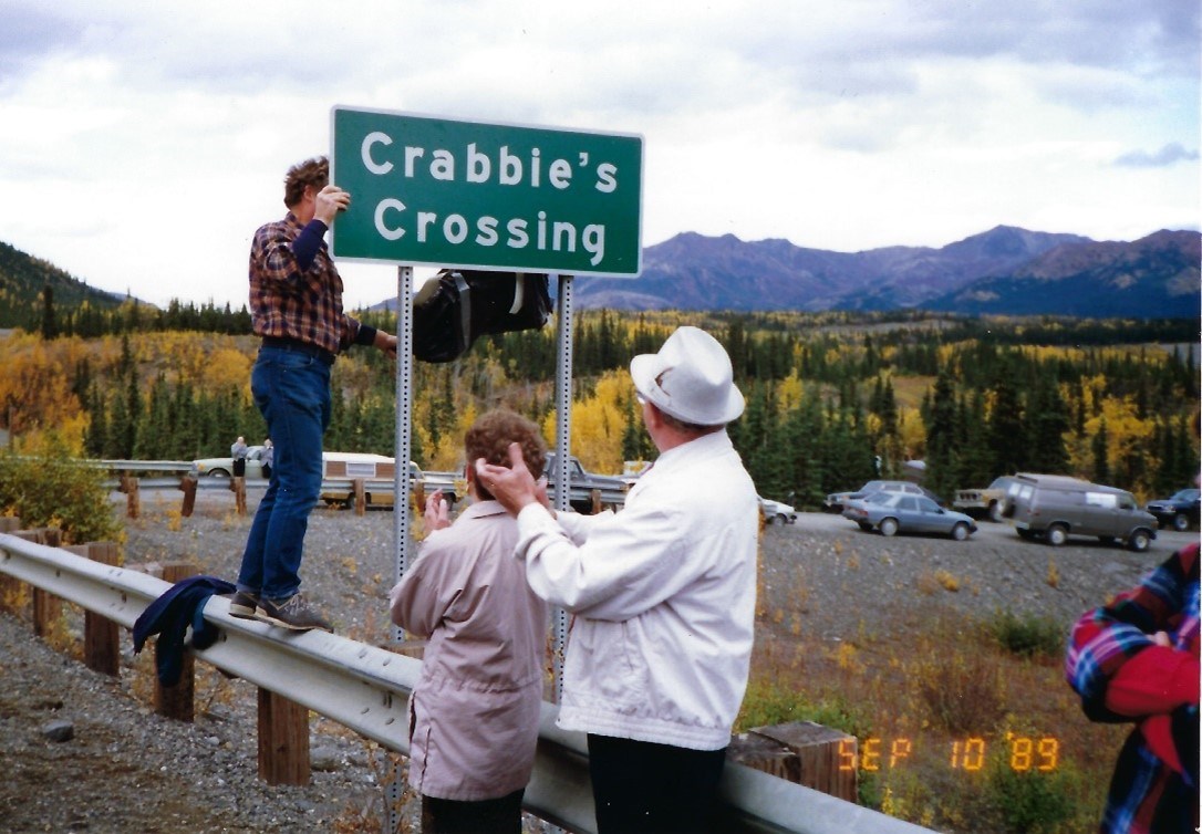 crabbies crossing 3 One person removes a plastic covering, revealing a green and white road sign that reads "Crabbie's Crossing". Two people stand on the road in front of the sign, clapping.