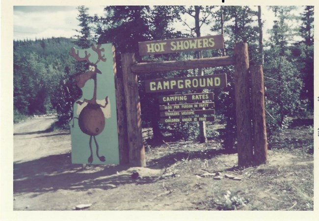 Wooden signs next to a dirt road read "Hot Showers, Campground, Camping Rates, $1.00 per person in party, trailers welcome, children under 12 free." A whimsical wooden moose decorates the left side of the sign.