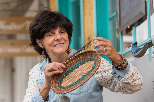 Woman holds and gazes at intricate, multicolored woven basket midway through creation process