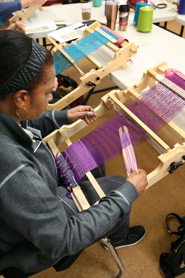 Seated weaver working at tabletop loom with purple thread.
