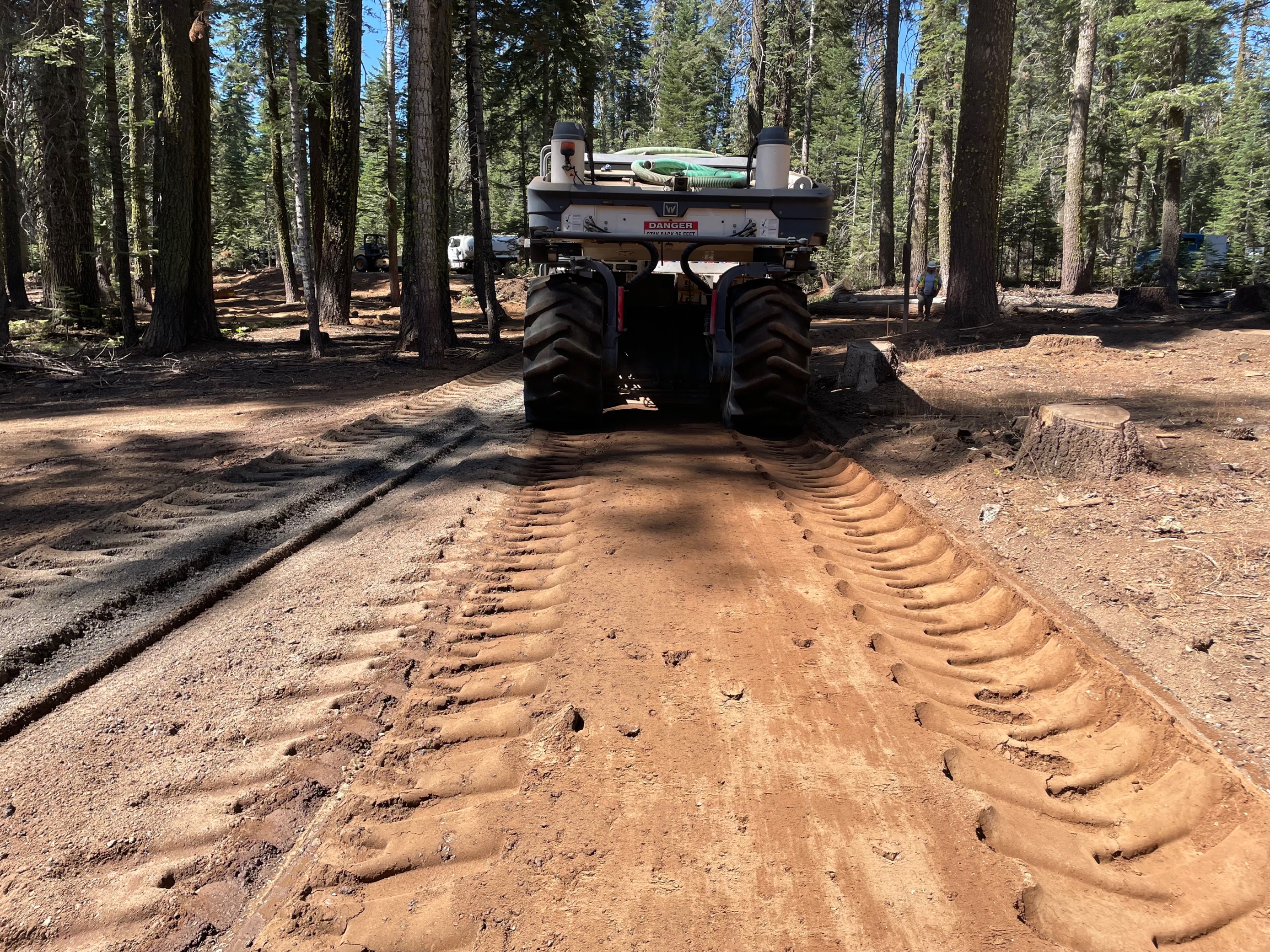 Large tire prints are left in the dirt road by a large construction vehicle in the forest.