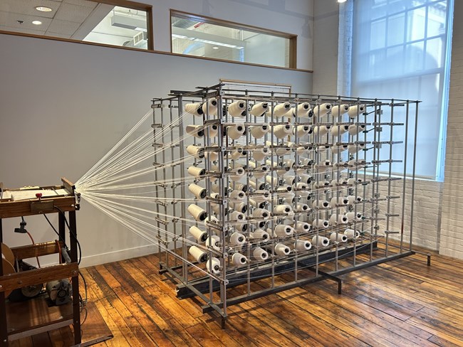 creel A metal frame full of many rolls of thread. Each roll has a thread that has been pulled forward into a machine.