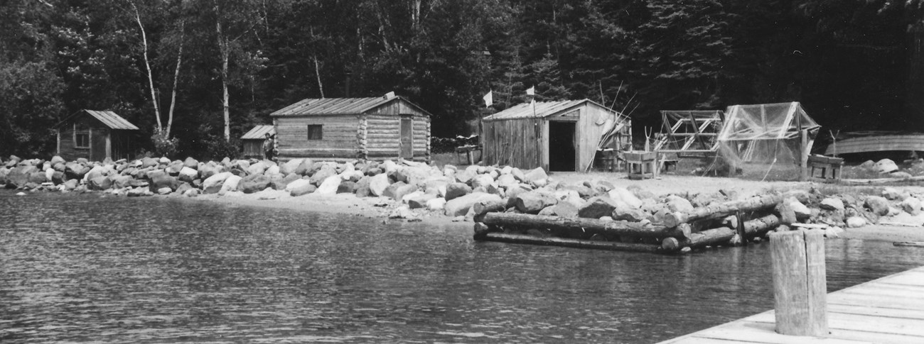 Black and white photo of historic wooden shacks in front of the forest, taken from the dock.