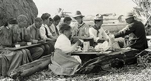 Group of men and women sit outdoors on logs around a makeshift table or with a plank across their laps eating dinner. The women wear long skirts.