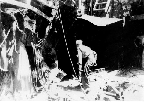 Black and white photograph of a ship's deck with warped metal and frayed cables. One sailor is crouching low in the center walking into a dark void in the ship.