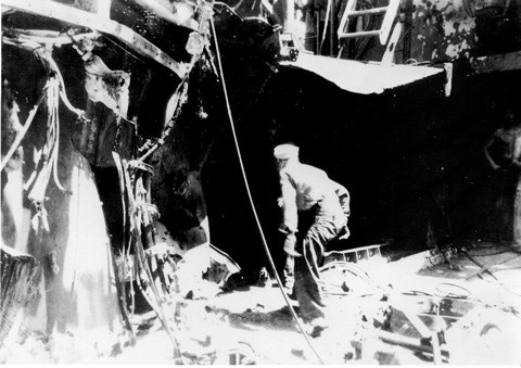 Damage to USS Cassin Young Black and white photograph of a ship's deck with warped metal and frayed cables. One sailor is crouching low in the center walking into a dark void in the ship.