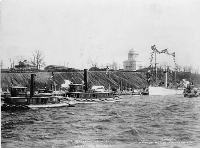 Black and white photo of a body of water with ships along the cliff shore and a white mausoleum at the top of the cliff