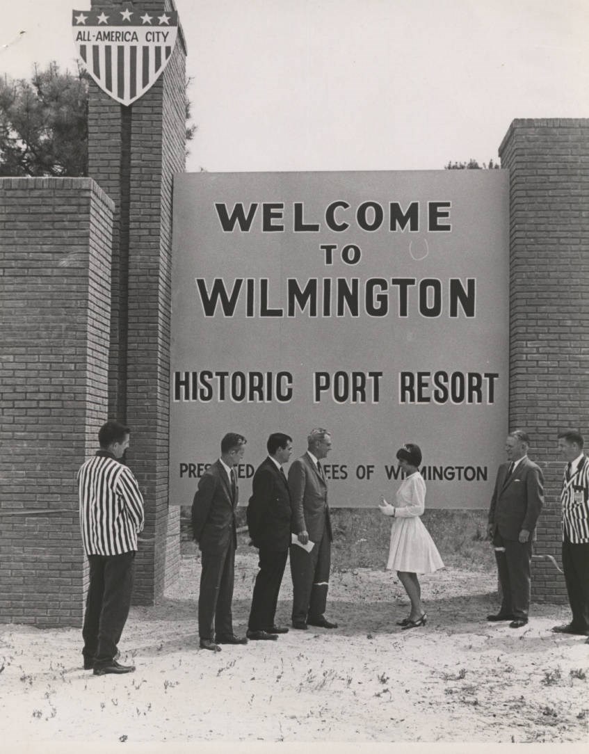 7 people standing in front of and looking at a "Welcome to Wilmington" sign about 30 feet tall. Sign in upper left corner designating Wilmington as an All-America City