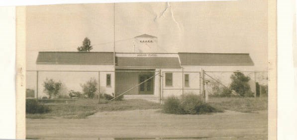 Yellowed black and white photograph of a long building with white walls and a pitched roof behind a tall chain link fence. A narrow tower extends above the recessed central section of the building and its entrance. There are few windows.