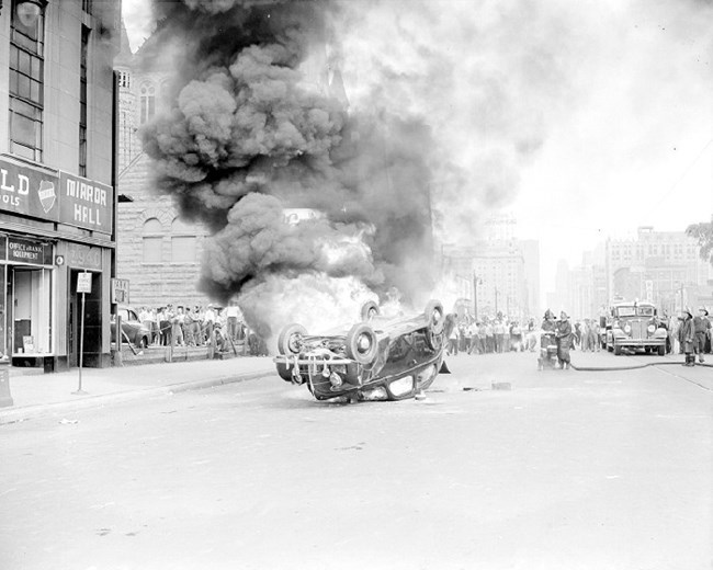 Smoke billows from an overturned car in the middle of a city street while firefighters attempt to extinguish it with a hose as a crowd looks on