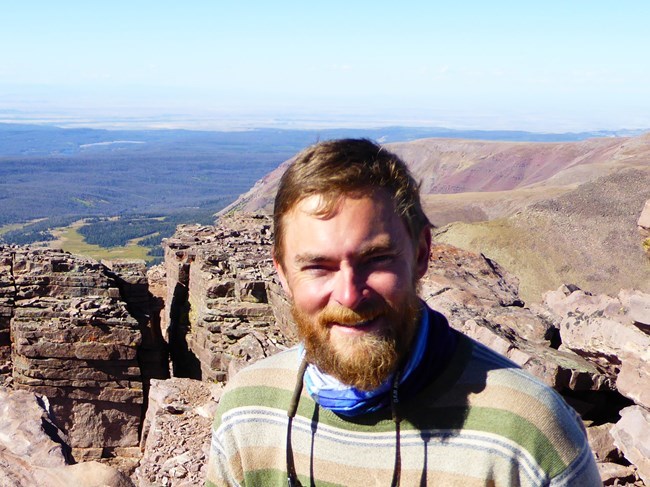 Headshot of man with beard smiling from a high rocky place with views of forests and meadows in distance.