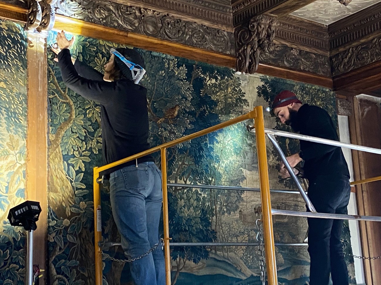 Two conservators on scaffolding carefully remove a tapestry from the wall.