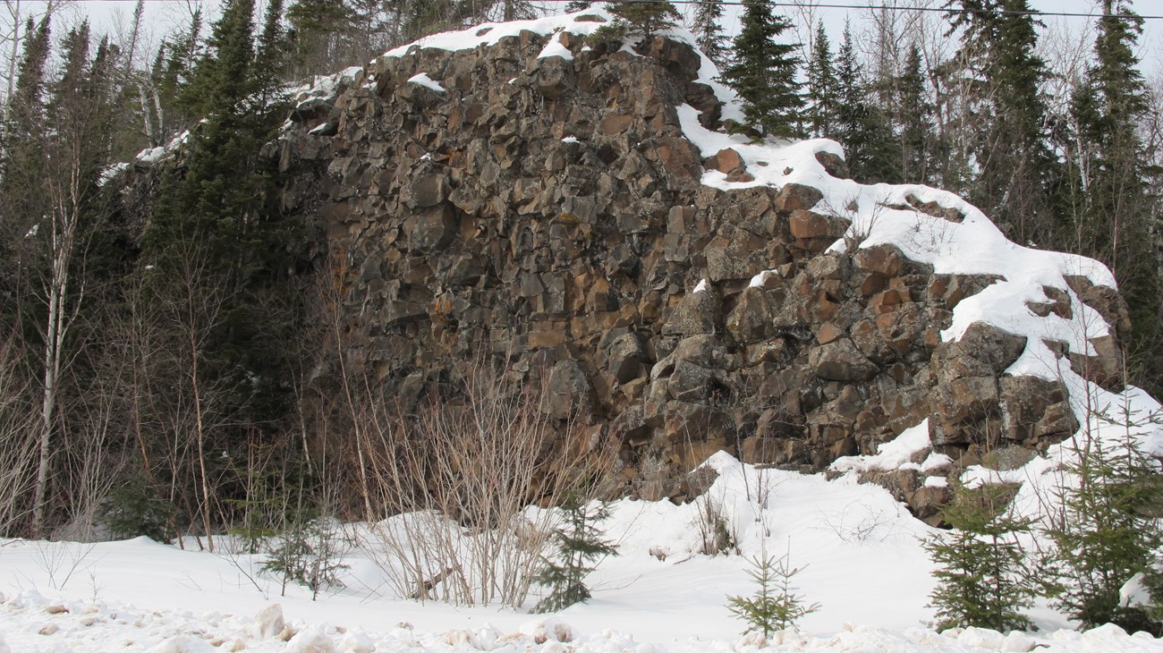 A natural dark rock wall with snow on top and in the foreground.