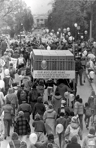 The Great Peace March for Nuclear Disarmament of 1986 arriving to Washington DC. November 14, 1986. black and white photo of a crowd marching to the capitol, carrying boxes with a sign that says "our nuclear home, 16,000 sticks of dynamite for every person on the planet.