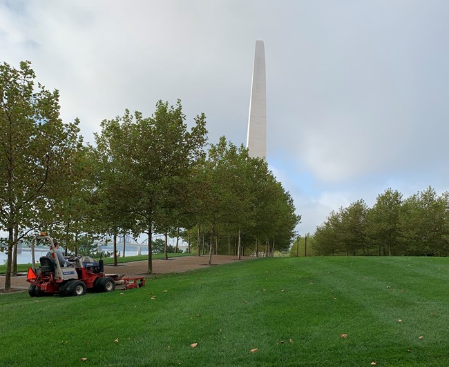 a woman on a riding lawn mower, with green grass and the Gateway Arch in the background.