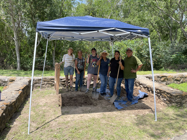 six people stand under a tent in front of a archeology unit on the ground