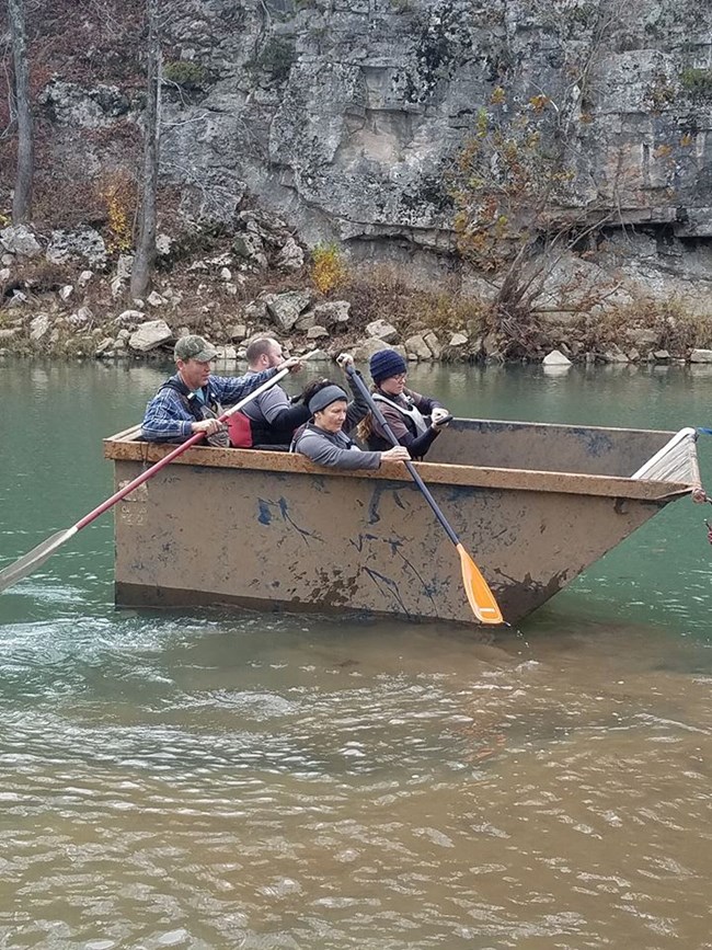 Four people paddle a floating dumpster down a river.