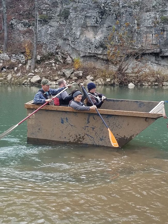 Dumpster Paddle Four people paddle a floating dumpster down a river.