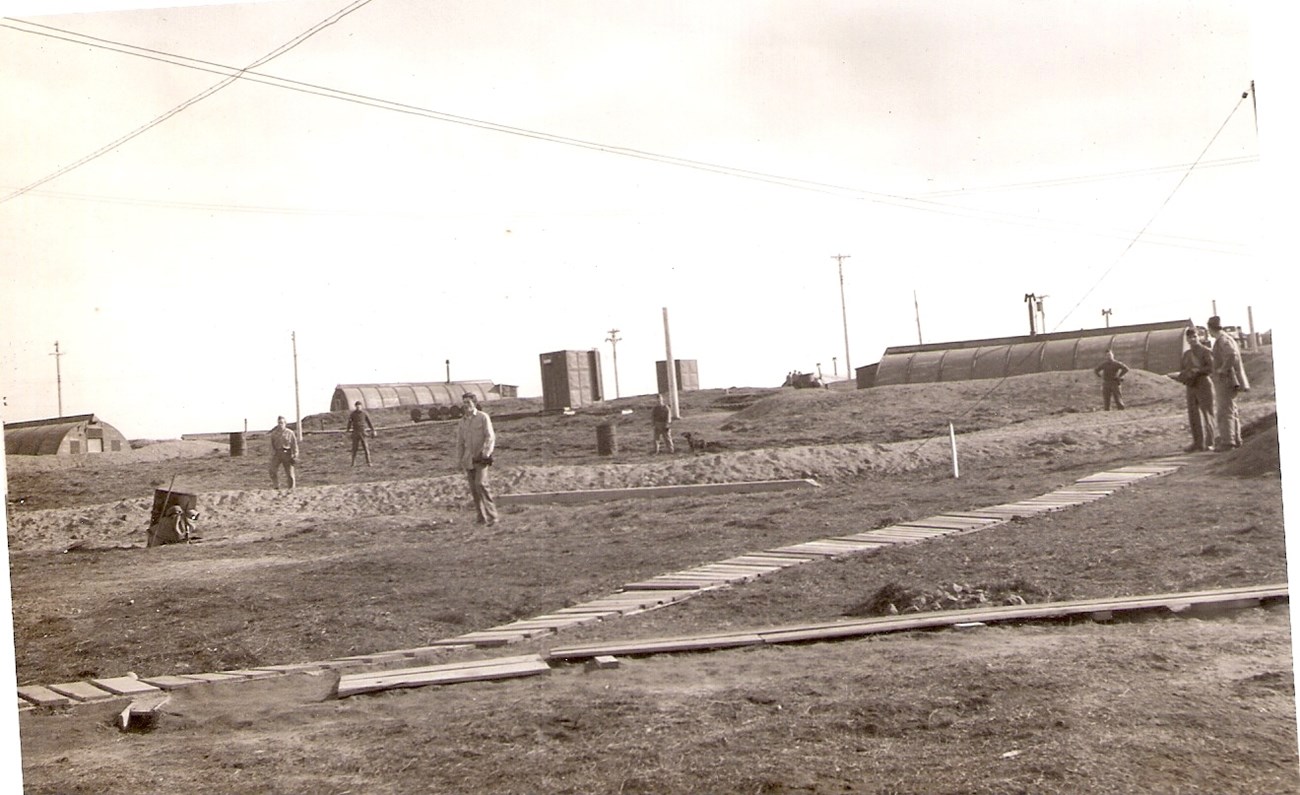 Dutch Harbor and 206th Coast Artillery Outside men scattered in grassy areas around Quonset huts and wooden boardwalks.