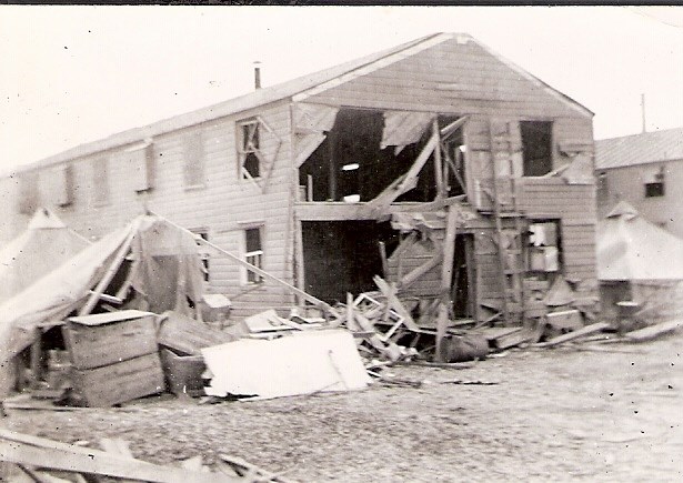 Two story wooden building with blown out walls and collapsed tents beside it.
