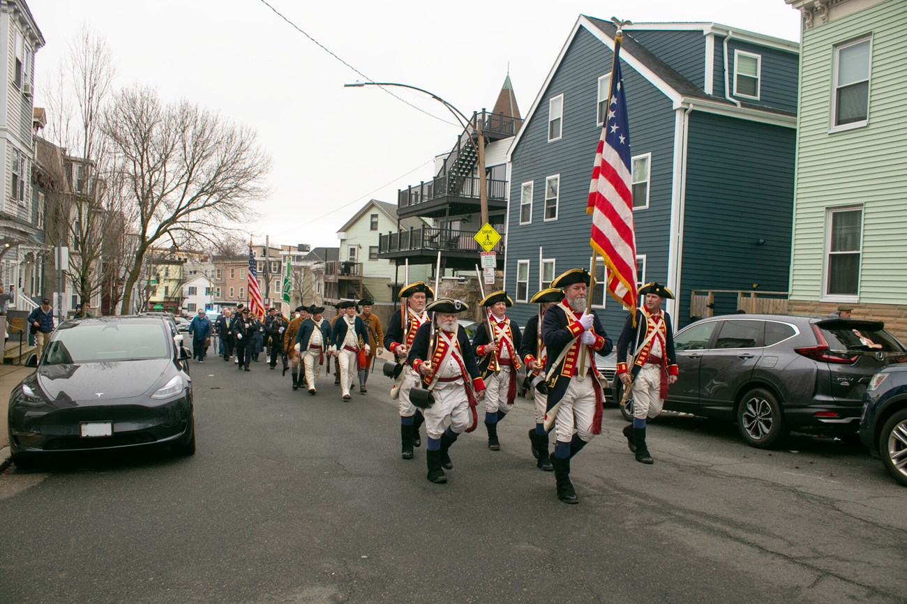 Minutemen dressed in 1700s kit walk in a procession down a street in South Boston.
