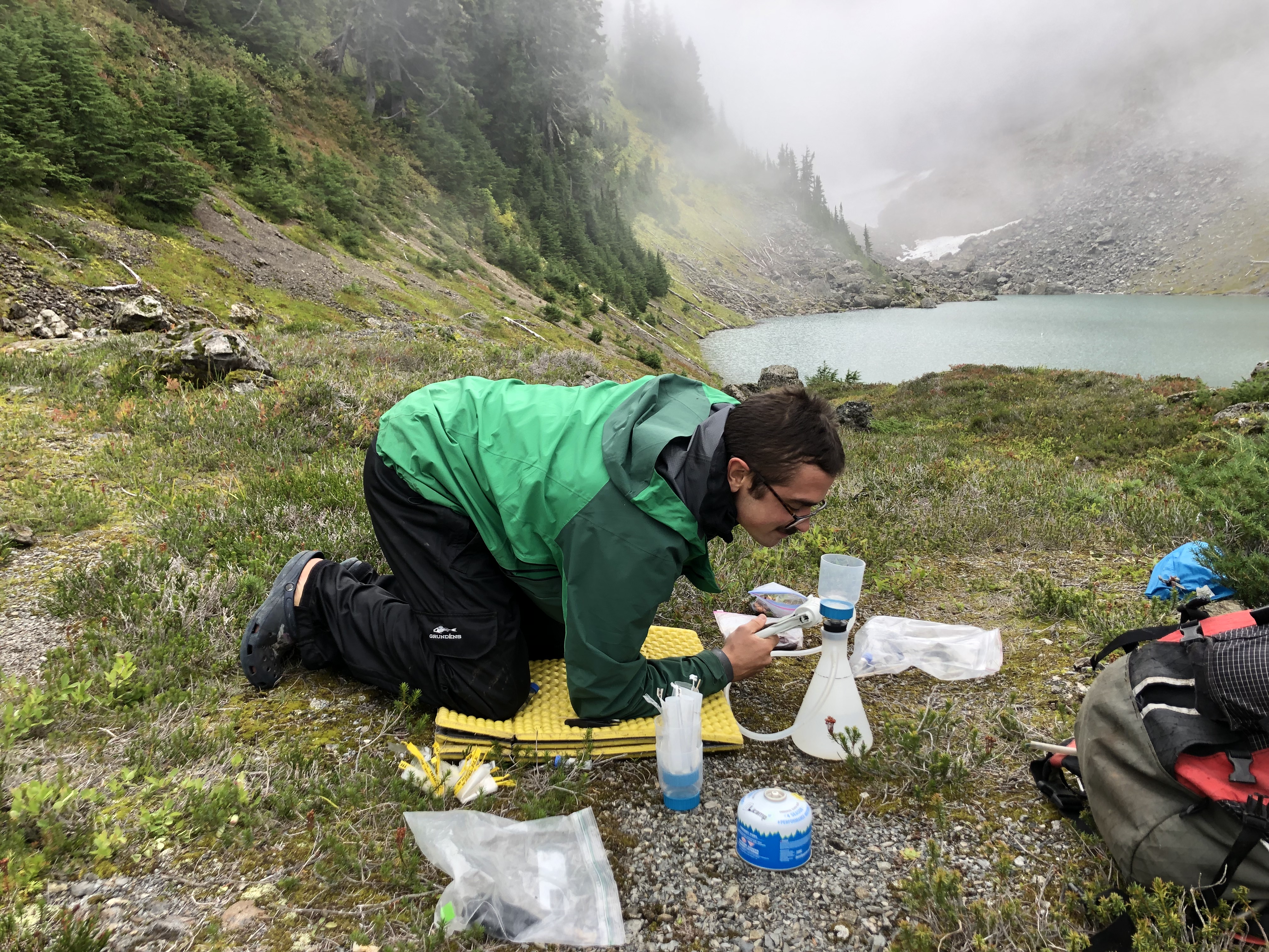 In front of a milky blue mountain lake, a person kneels on a foam pad holding a hand pump attached to a plastic flask.