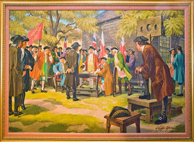 men in colonial dress standing, with flags, table and wooden building visible