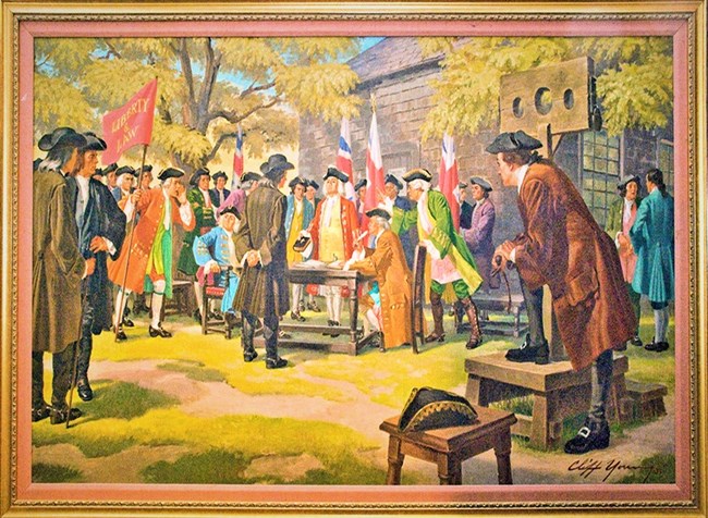 Many men, in colonial clothes, standing near a table, with one of them arms folded behind his back, and another man extending a book toward him, with a wooden building in the background