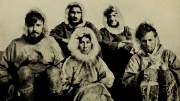 A black and white image of the Wrangel Island expedition party seated wearing fur coats. Ada Blackjack is seated in the middle of the group, and Allan Crawford pulls a string to the left to operate the camera.