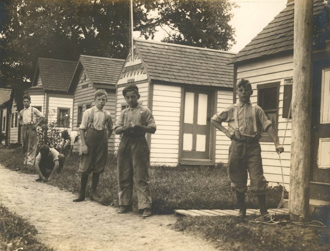 Six boys stand along a small dirt path in front of four cottages. Two boys are crouched over, the others stand with hands on their hips.