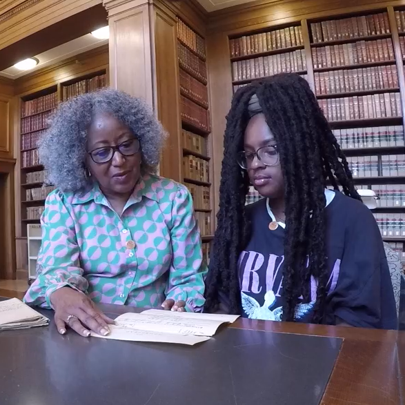 fb6aa5f8-0d2d-42d4-91bd-d2a8bc06b143 A woman shows a young adult a document in a library setting.