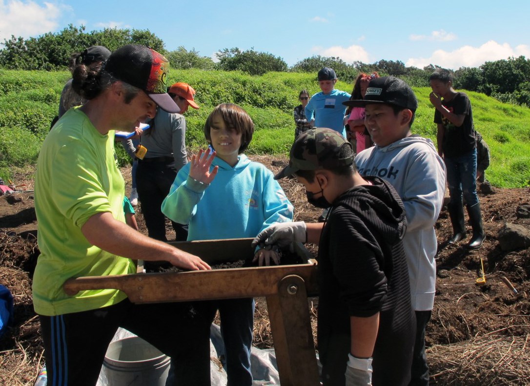 Archeologist shows three students how to sift dirt to find potential artifacts from excavation.