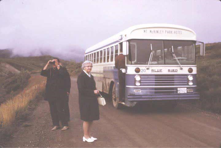 two women standing outside of a blue and white bus with marquee reading mount mckinley park hotel, on a dirt road