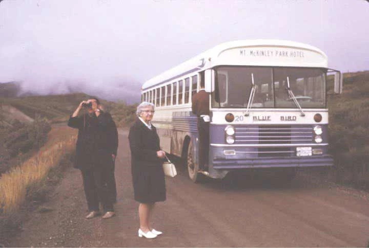 fig-1 two women standing outside of a blue and white bus with marquee reading mount mckinley park hotel, on a dirt road