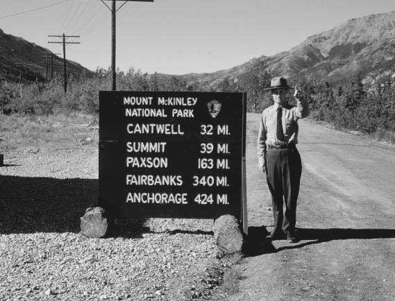 uniformed park ranger standing next ot a sign that reads mount mckinley national park and mileages to locations in alaska