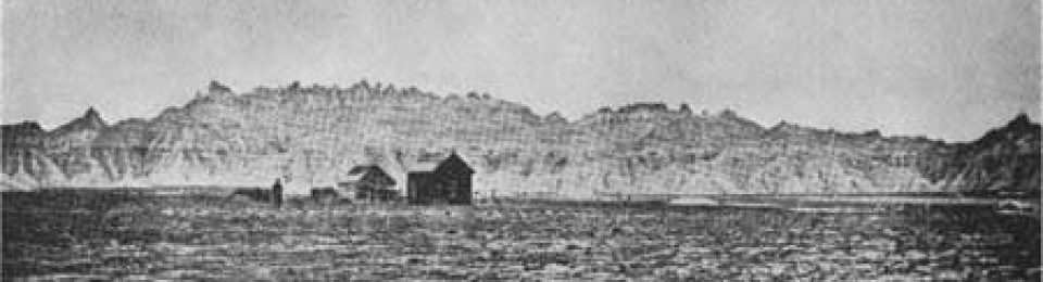 a historic black and white photograph shows a ramshackle house sitting in front of jagged badlands formations