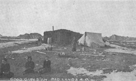 lt= a historic black and white photograph in which three women sit in front of a basic home with the caption "good girls in bad lands SD"