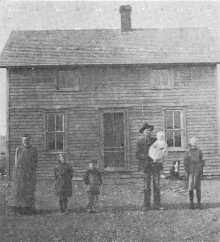 a family of six stands in front of a two-story wooden home in a historic black and white photograph