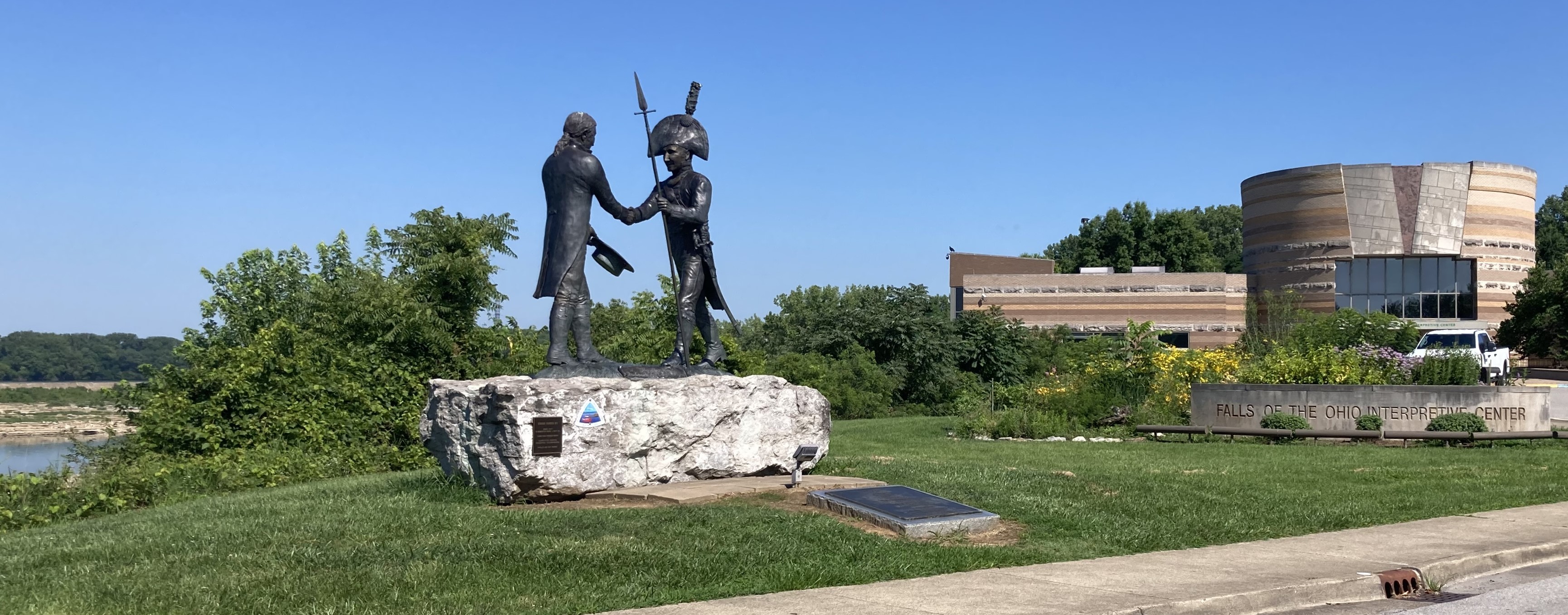 A landscape with a statue of two men shaking hands on a rock on the left and a building on the right built of different colored rocks, giving a banded appearance. Rock outcrops are present in the background on the left.