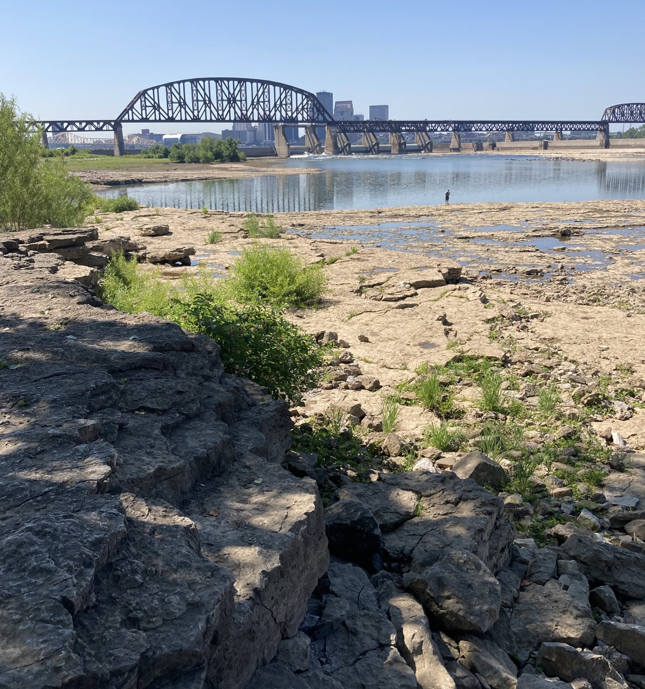 A landscape with rock outcrops near water level in the foreground and a long bridge with two sets of trusses in the background.